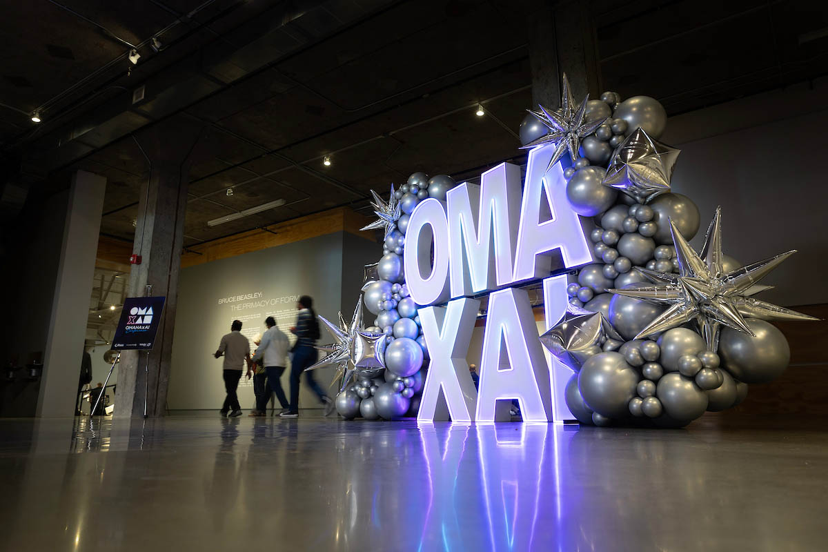Large illuminated "OMA x AI" letters surrounded by silver balloon sculptures in an event lobby area.